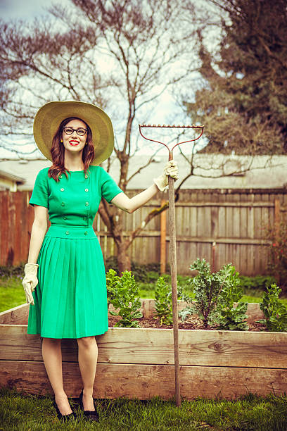 A young woman in 1950s style pleated dress, high heels, glasses, bright red lipstick and wearing a large floppy straw sun hat takes a break from working on the yard and garden, smiling with her work gloves and rake in hand.  Vertical with copy space.