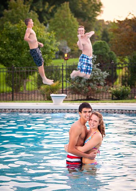 Summertime shenanigans and relationships Two mischievious teen boys launched off the diving board as another teen couple poses for a casual portrait. They're about to get splashed and surprised. photo bomb stock pictures, royalty-free photos & images