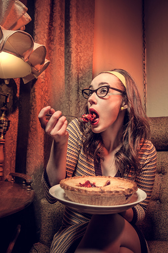 A woman in 1960s style clothes and living room sits in an arm chair and eats a whole homemade pie by herself, a bored, tired expression on her face. The woman wears cats eye glasses, bright red lipstick and a 60's style headband.  Orange tinted light and brown living room colors.  Vertical with copy space.