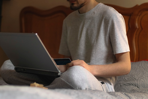 A young caucasian man in gray sweatpants and a t-shirt holds a bank card in his hand and type on the keyboard of a laptop that he holds on his knees, and next to it is a small kraft gift box, sitting on the bed in the bedroom, close-up side view with depth of field.Online shopping concept, at home, lifestyle, business, finance, gifts.