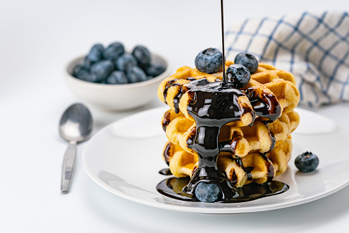 Pouring chocolate syrup on stack of fresh delicious homemade waffles topping with ripe organic blueberry fruit on white ceramic dish with blueberry fruit in white ceramic cup and metal spoon on white background.