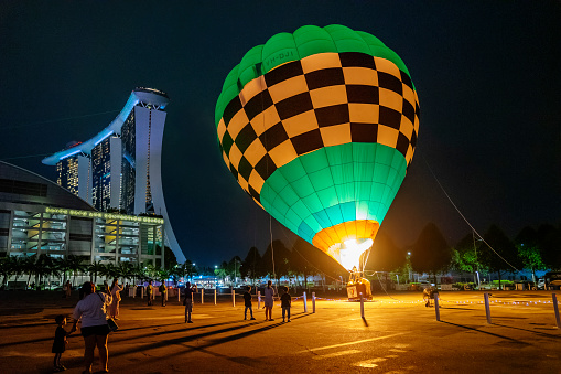 Singapore, Singapore - October 8, 2022: Spectators watch as a tethered hot air balloon operated by Ballons du Monde is set up at an open space beside Marina Bay Sands and its convention centre at night.