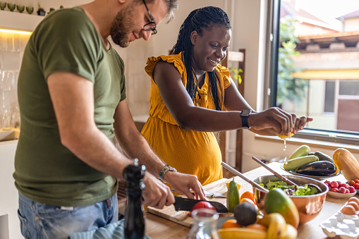 Photo of happy black pregnant woman preparing healthy food in the kitchen with her husband.