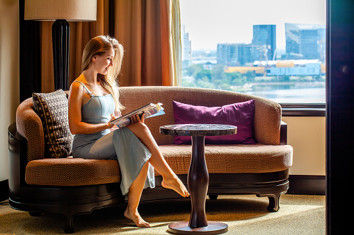 Business woman sitting on couch, reading magazine, looking at city view from window in modern luxury hotel room. Taking rest after hard working day. Young businesswoman in suit relaxing on weekend.