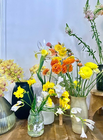Vertical close up of bright spring interior flowers of yellow, white and orange tones in vases on wood table against white wall