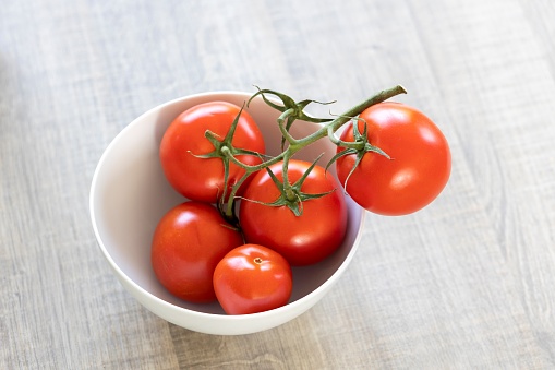 A portrait of a white bowl with five delicious red tomatoes which are still on the vine. The healthy and fresh vegetables have a different size and are ready to be used in the kitchen for cooking