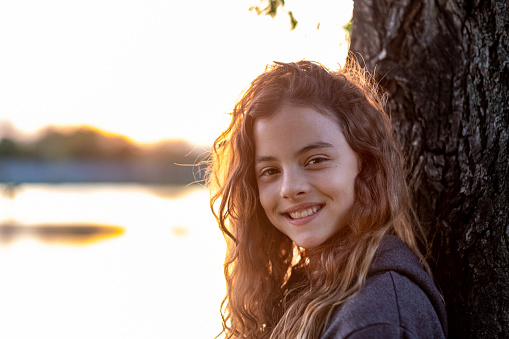 Portrait smiling girl standing near tree trunk at riverbank