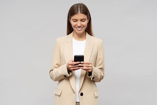 Young attractive female CEO in jacket using smartphone isolated on gray  background to make appointment with partners to discuss new service supply for next week. Business and technology