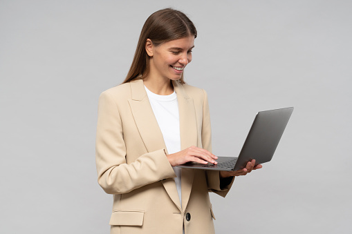 Portrait of female office manager in jacket working on laptop online standing on gray background. Pretty secretary checking e-mail, booking plane tickets for business trip of her ceo