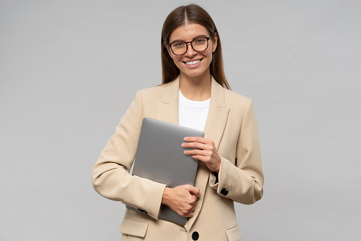 Studio portrait of happy young female teacher in glasses and jacket with laptop in hands after classes standing on gray background smiling. Modern education and technology