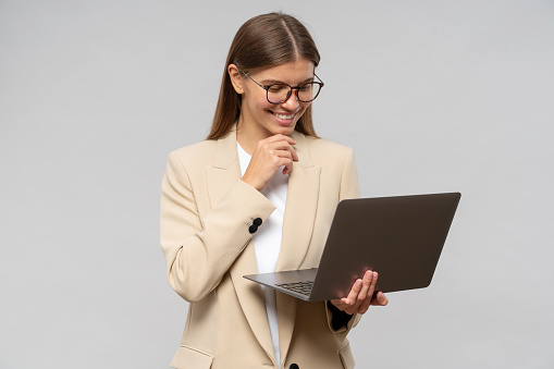 Portrait of happy female teacher in glasses and jacket having lesson online on laptop, listening to her student attentively with interest, touching chin with hand on gray studio background