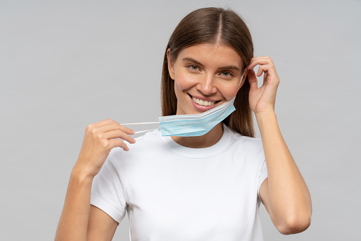 Pretty happy woman taking medical mask off on gray background celebrating the end of coronovirus pandemic, lock down and quarantine, smiling at camera wearing white t-shirt with copy space