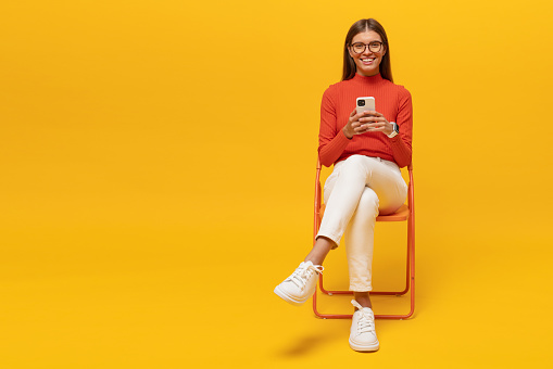 Pretty young woman enjoying spending leisure time surfing mobile internet sitting on chair on yellow copy space background with smartphone in hands, looking at camera smiling, reading funny memes