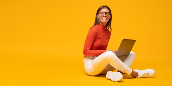 Side view of woman in glasses and stylish clothes spending leisure time sitting on floor using laptop, watching favorite online show or podcast on yellow background with copy space on the right