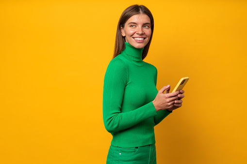 Side view portrait of attractive woman blogger sharing post in her business account in social media network looking aside on yellow background. Smiling smm specialist working on smartphone