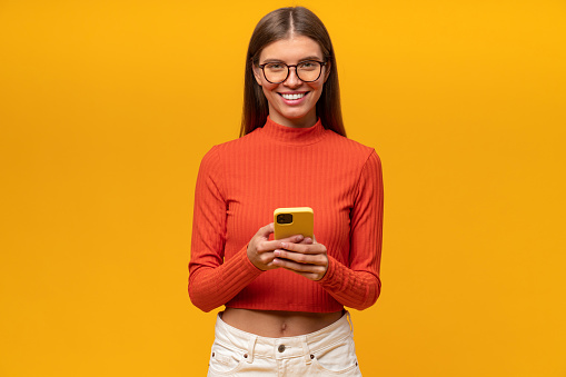 Pretty happy smiling student girl in casual clothes and glasses using studying app to learn foreign language, watching webinar, surfing mobile internet, browsing useful web pages