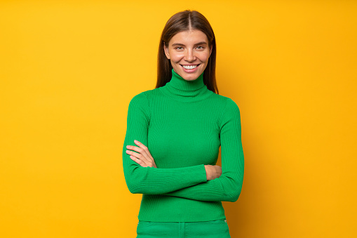 Portrait of stylish creative fashion designer or editor of popular magazine in green turtleneck standing in bossy posture with crossed hands on yellow background with happy smile