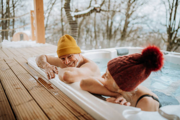 Senior couple in kintted cap enjoying together outdoor bathtub at their terrace during cold winter day. stock photo