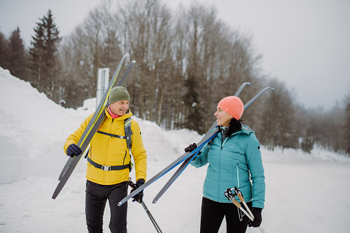 Senior couple crossing winter forest with skis on shoulders.