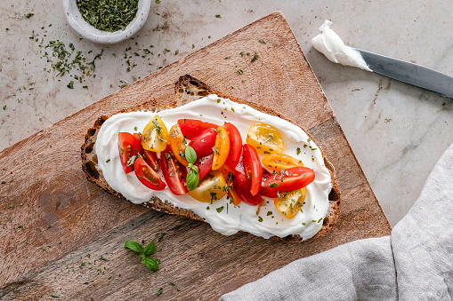 Overhead view of a toasted slice of sourdough bread topped with cream cheese, tomatoes and herbs, served on a wooden board