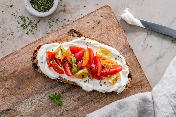 Overhead view of a toasted slice of sourdough bread topped with cream cheese, tomatoes and herbs, served on a wooden board