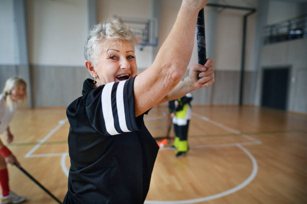 Excited senior woman playing floorball in gym with her friends. stock photo