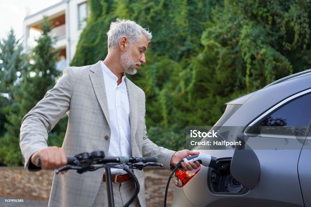 Close up of businessman in suit on way to work standing at eletric scooter and charging his electric car. Concept of eco commuting and green transportation. Close up of businessman in suit on way to work standing at an eletric scooter and charging his electric car. Concept of eco commuting and green transportation. Car Stock Photo