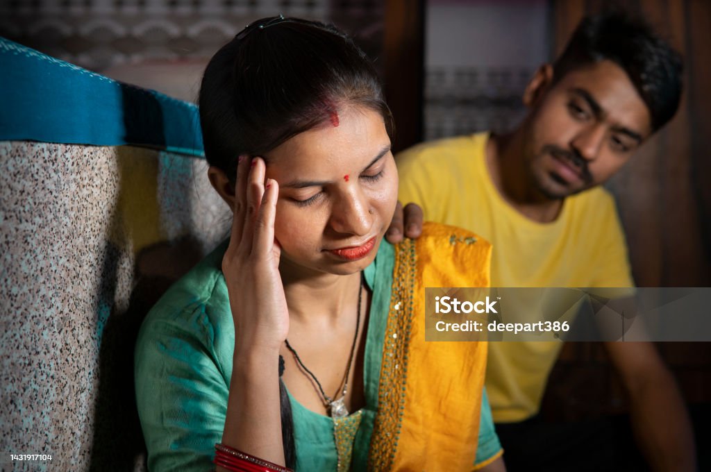 Indian man consoling his wife who is thinking about problems by holding her head at home. Indoor image of a sad, depressed married young Indian woman holding her head out of headache while thinking about family and relationship problems and her husband consoling her with a hand on her shoulder from behind at home. She is in traditional clothes Salwar Kameez and dupatta. Couple - Relationship Stock Photo