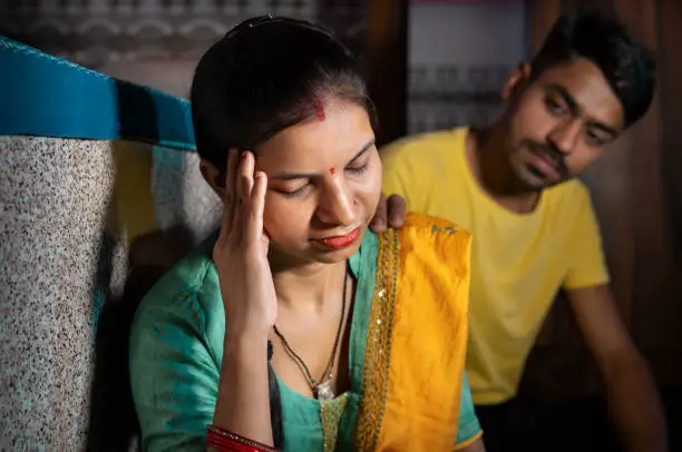 Indoor image of a sad, depressed married young Indian woman holding her head out of headache while thinking about family and relationship problems and her husband consoling her with a hand on her shoulder from behind at home. She is in traditional clothes Salwar Kameez and dupatta.
