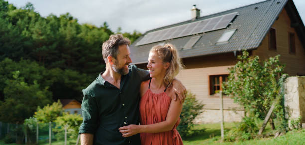 Happy couple standying near their house with solar panels. Alternative energy, saving resources and sustainable lifestyle concept. stock photo