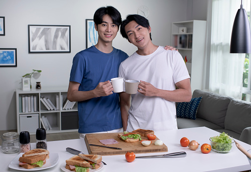 Portrait of romantic Asian gay couple cooking on kitchen and looking at camera. Lgbt men couple are having fun together while preparing healthy food. Healthy lifestyle concept.