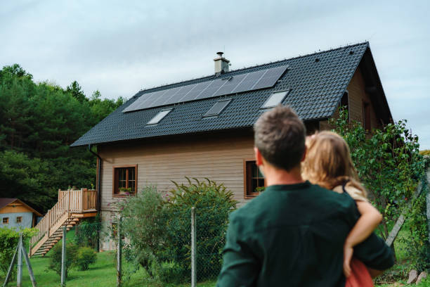 Rear view of dad holding her little girl in arms and looking at their house with installed solar panels. Alternative energy, saving resources and sustainable lifestyle concept. stock photo