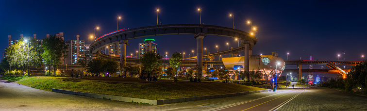 People relaxing in the popular Ttukseom Hangang Park at night beneath the street lights of Cheongdam Bridge and the high-rise cityscape panorama of downtown Seoul beside the Han River in South Korea.