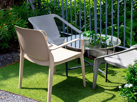 Three empty beige plastic chairs with round glass table with small plant pot in the outdoor garden on artificial grass floor and grey iron fence. Table set relaxing in garden.