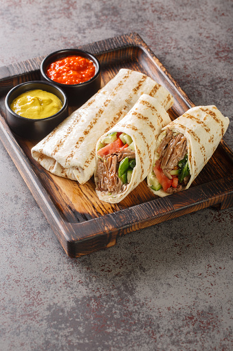 Doner kebab made of meat cooked on a vertical rotisserie with additions of vegetables and sauce wrapped in lavash closeup on the wooden board on the table. Vertical