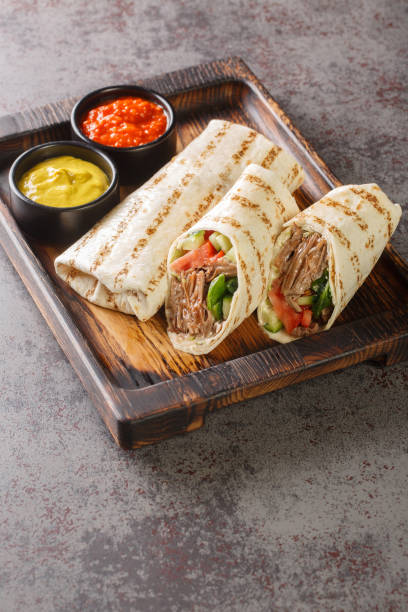 Doner kebab made of meat cooked on a vertical rotisserie with additions of vegetables and sauce wrapped in lavash closeup on the wooden board. Vertical stock photo