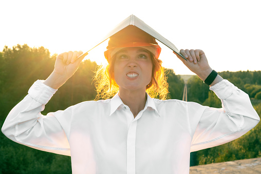 A female inspector in a red construction helmet in a white shirt makes a face and holds a tablet with a house. The concept of an incompetent employee.