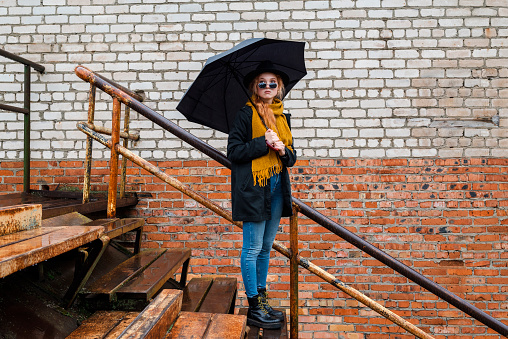 Woman in a black round felt hat and black sunglasses the background of a brick city wall. Girl in a warm orange scarf and coat looks out from under her glasses holding an umbrella in her hand.
