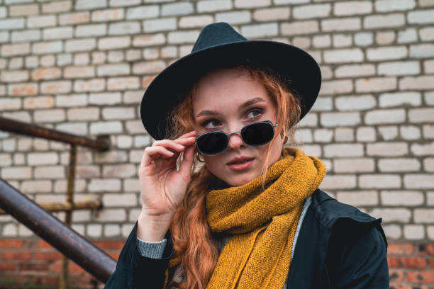 Portrait of a woman in a black round felt hat and black sunglasses against the background of a brick city wall. A girl in a warm orange scarf and coat looks out from under her glasses.
