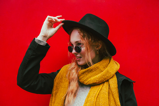 Portrait of a smiling happy girl in a black round felt hat and sunglasses with an orange scarf. Female autumn city portrait on the background of a red steel wall.