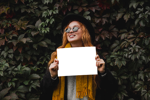 An outdoor girl in autumn clothes holds an empty sheet of paper in her hands while standing against a background of foliage. A woman with a mock up.