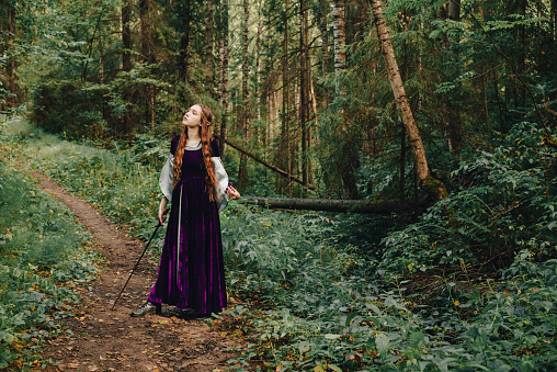 An elven girl driver with long red hair in a purple dress in the forest poses standing with a medieval sword. The woman is a fairy tale elf character.