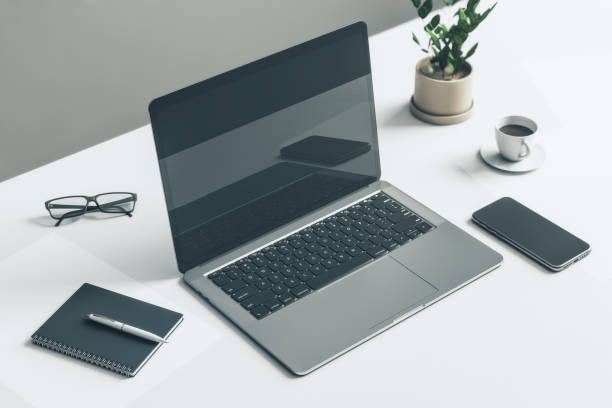 istock Top side view on blank dark modern laptop screen with reflection on white table with glasses, notebook, smartphone and flowerpot on grey background. 3D rendering, mockup 1431981926