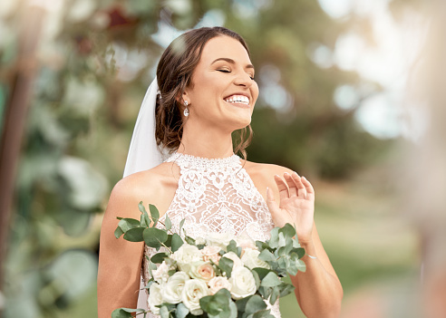 Excited bride in wedding with bouquet in nature park with green trees, bokeh and summer sunshine. Happiness, commitment and dream of a beauty woman with flowers for marriage in outdoor lens flare