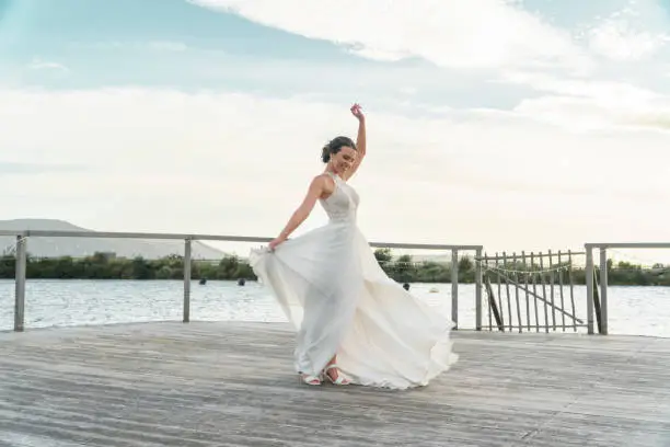 Photo of Bride with white wedding dress, happy to pose outdoor on a jetty with lake or dam in background. Woman smile on day of marriage celebration outside with water, trees and mountain in nature backdrop