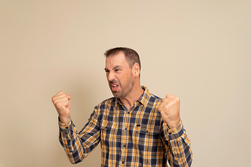 Handsome hispanic man with beard wearing checkered shirt over isolated beige background angry and crazy raising fists frustrated and furious while shouting with anger. Rage and aggressive concept.