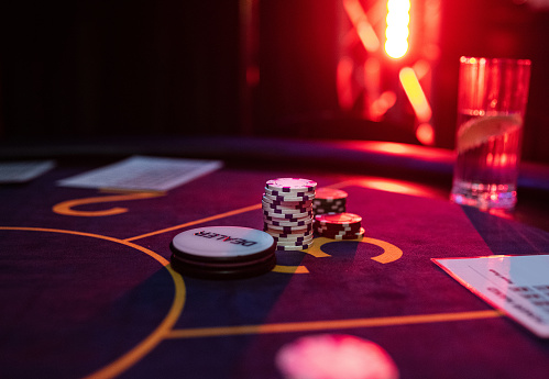 a stack of poker chips in a casino on a table with a dealer's mark nearby, a cozy, dark atmosphere, a glass of water with lemon nearby, red light on the background. casino chips on gambling table.  Background for the gaming business. Winner, fun, wealth concept.
