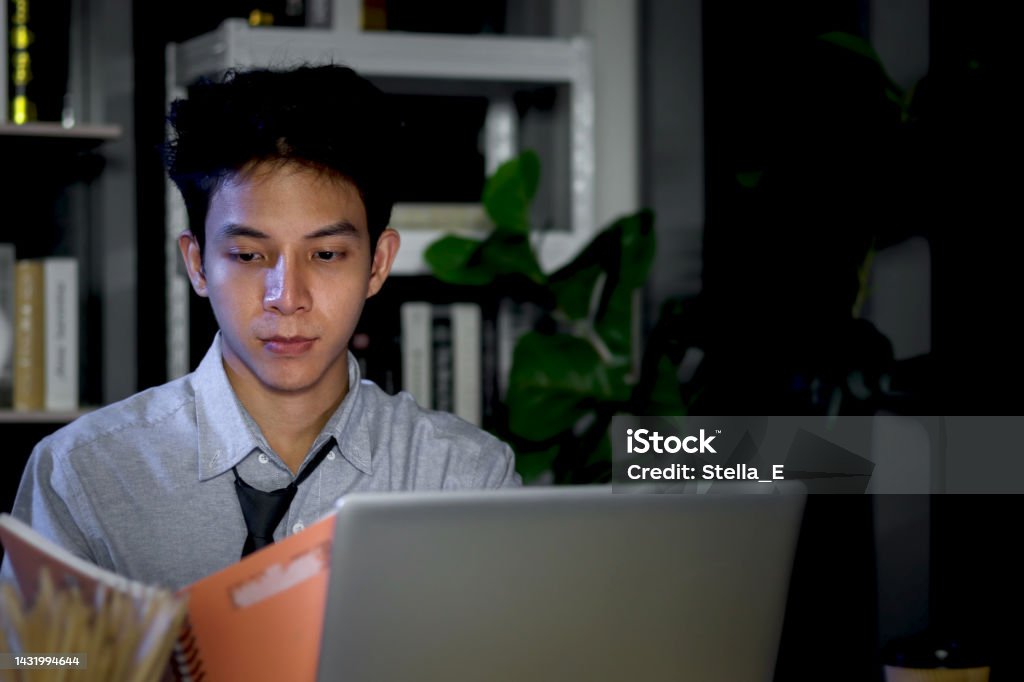 Asian young tired staff officer man using laptop computer having overwork project overnight in office, exhausted unhappy businessman feeling sleepy after after working hard overtime at night. 20-24 Years Stock Photo
