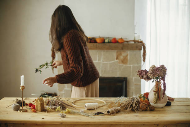 istock Making autumn wreath on rustic table at fireplace. Stylish woman arranging dried grass in wreath on wooden table with scissors, thread, pumpkin, candle. Fall decor and arrangement in farmhouse 1431998069