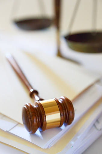 A gavel, photographed with a very shallow depth of field, rests on top of a stack of file folders and legal documents. A justice scale sits out of focus in the background.